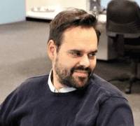 A person with short hair and a beard is sitting in an office environment, turned slightly to the side. The person is wearing a dark sweater over a collared shirt. Office furniture and equipment are visible in the background.